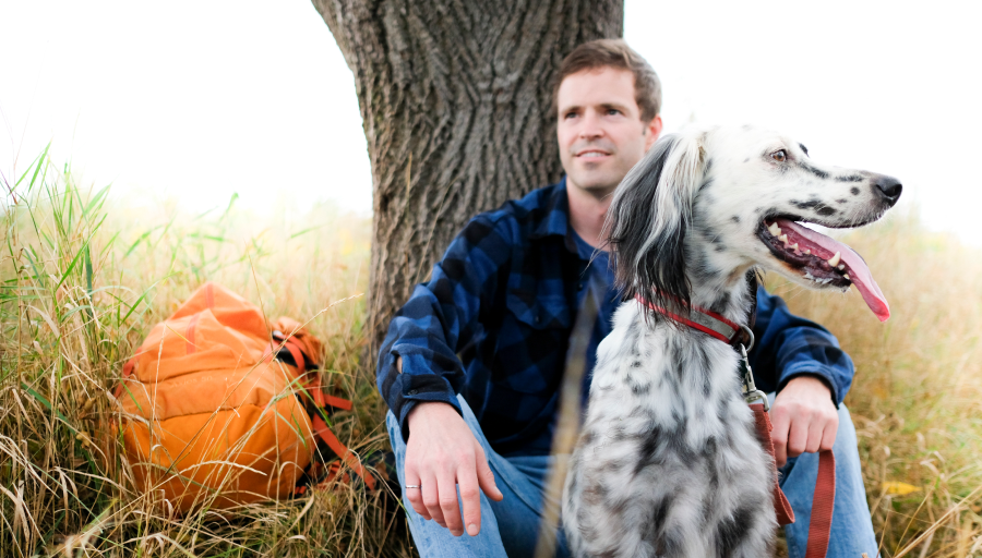 Man sat in front of a tree with his orange backpack on the grass and his dog sat in front of him on a leave
